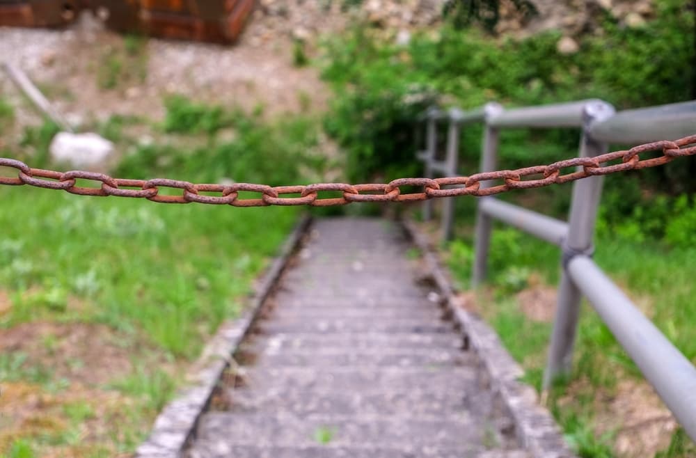 Old, rusty chain in front of a stone staircase with a metal railing and some overgrown vegetation
